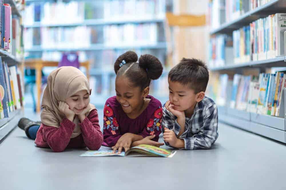 Three (3) kids who are friends are laying on the floor in between bookshelves in the library. They are reading a book together. They are all smiling and enjoying their afternoon.