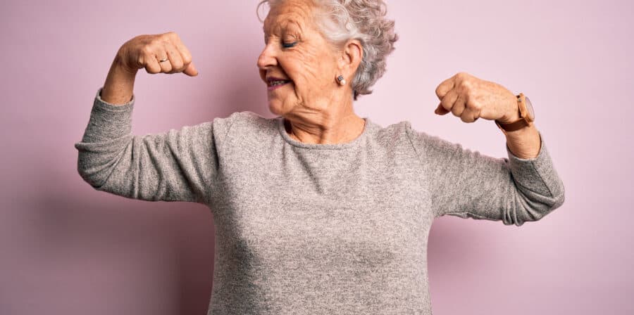 Senior beautiful woman wearing casual t-shirt standing over isolated pink background showing arms muscles smiling proud. Fitness concept.