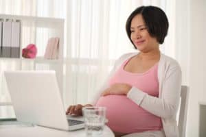Waist-up portrait of smiling Asian pregnant woman using modern laptop while working from home, cozy interior of living room on background
