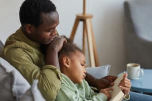 Side view portrait of loving African-American father and daughter reading while sitting on couch together in cozy home interior, copy space