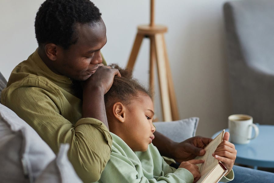 Side view portrait of loving African-American father and daughter reading while sitting on couch together in cozy home interior, copy space