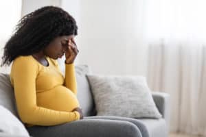Young pregnant black woman suffering from headache or migraine, feeling sick, sitting on sofa at home, free space. Sad african american expecting lady touching her forehead and big tummy, home alone