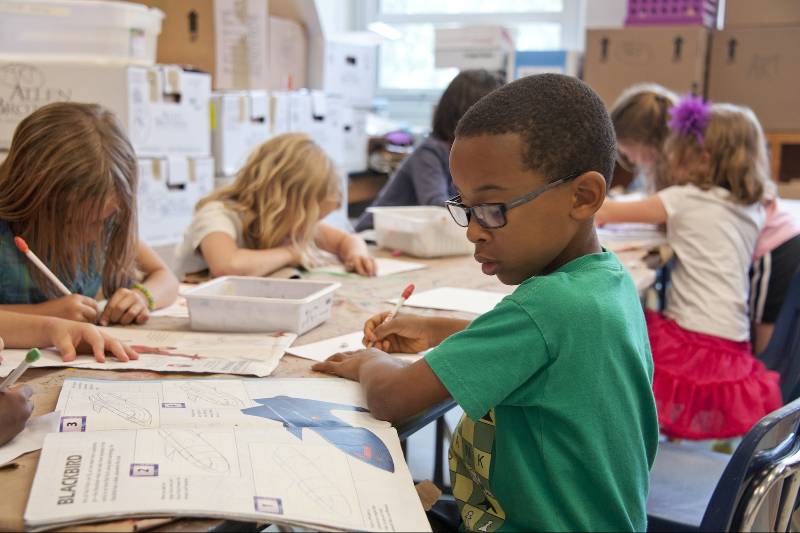 Captured in a metropolitan Atlanta, Georgia primary school, seated amongst his classmates, this photograph depicts a young African-American schoolboy who was in the process of drawing with a pencil on a piece of white paper. Note that the student was focused on a drawing book that referenced fantasy flying planes, while intent on creating his artwork, seemingly oblivious to all the classroom goings-on that surrounded him. It is important to know that objects, including pencils, crayons, paper, etc., are known as fomites, and can act as transmitters of illnesses.