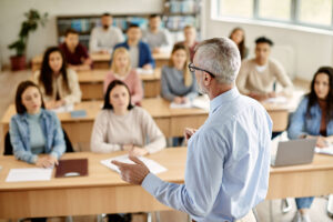 Back view of mature professor giving lecture to large group of college students in the classroom.