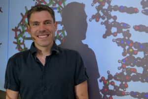 Sascha Duttke, an assistant professor in the School of Molecular Biosciences in Washington State University's College of Veterinary Medicine, poses for a photo with a projected image of DNA strands (photo by College of Veterinary Medicine/Ted S. Warren).