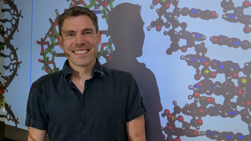 Sascha Duttke, an assistant professor in the School of Molecular Biosciences in Washington State University's College of Veterinary Medicine, poses for a photo with a projected image of DNA strands (photo by College of Veterinary Medicine/Ted S. Warren).