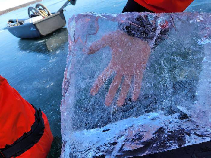 A hand can be seen through a clear block of black lake ice