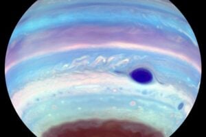 An artificially colored ultraviolet image of Jupiter reveals the Great Red Spot as a striking blue feature, while an oval of concentrated haze appears in the brown haze near the south pole. This dark oval, likely caused by mixing from a high-altitude vortex, mirrors similar but less frequent features seen at the planet's north pole.