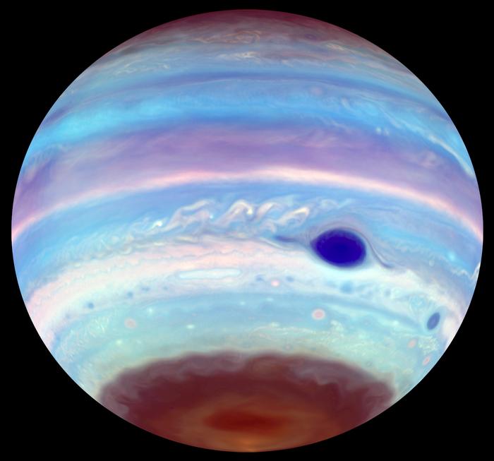 An artificially colored ultraviolet image of Jupiter reveals the Great Red Spot as a striking blue feature, while an oval of concentrated haze appears in the brown haze near the south pole. This dark oval, likely caused by mixing from a high-altitude vortex, mirrors similar but less frequent features seen at the planet's north pole.