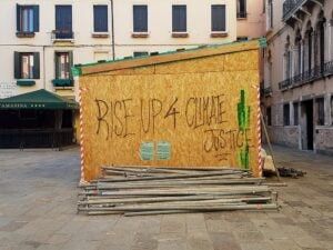 A wooden construction panel in a Venetian square, Campo Santa Marina, features black spray-painted graffiti reading ‘RISE UP 4 CLIMATE JUSTICE.’ The panel is part of a temporary structure, bordered by red and white hazard tape, with a pile of metal pipes stacked on the cobbled ground in front. Behind, historic buildings with shuttered windows and balconies frame the scene.