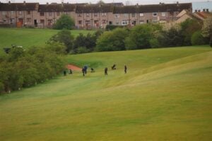 Several golfers play on the 10th fairway and green of Canmore Golf Club in Dunfermline, Scotland, with a row of terraced houses visible in the background and a line of trees separating the golf course from the residential area.