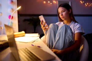 Worried Teenage Girl Sitting At Desk In Bedroom At Home Looking At Mobile Phone At Night