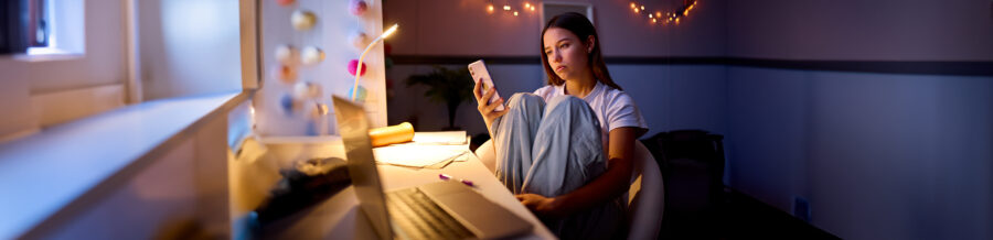 Worried Teenage Girl Sitting At Desk In Bedroom At Home Looking At Mobile Phone At Night