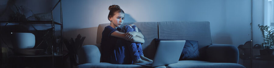Frightened teenage girl with laptop on sofa in dark room. Danger of internet
