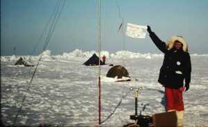 A person in heavy cold-weather gear holds up a handmade sign at a temporary research camp on the Arctic ice near the North Pole. The snow-covered scene includes several pitched tents, icy ridges in the background, scientific instruments in the foreground, and clear blue skies.