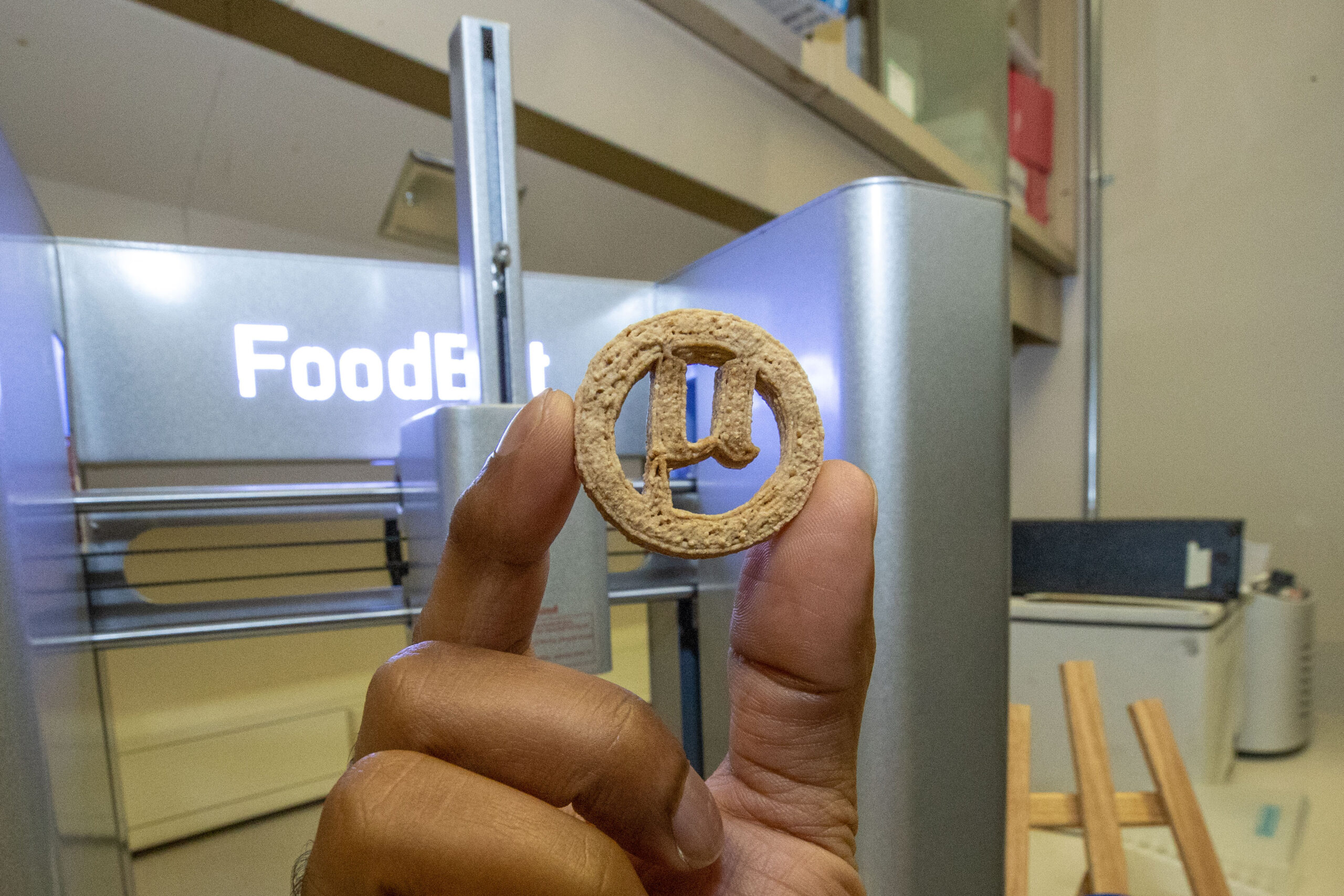 Hand holding a mu-shaped µBites cookie in front of a 3D printer