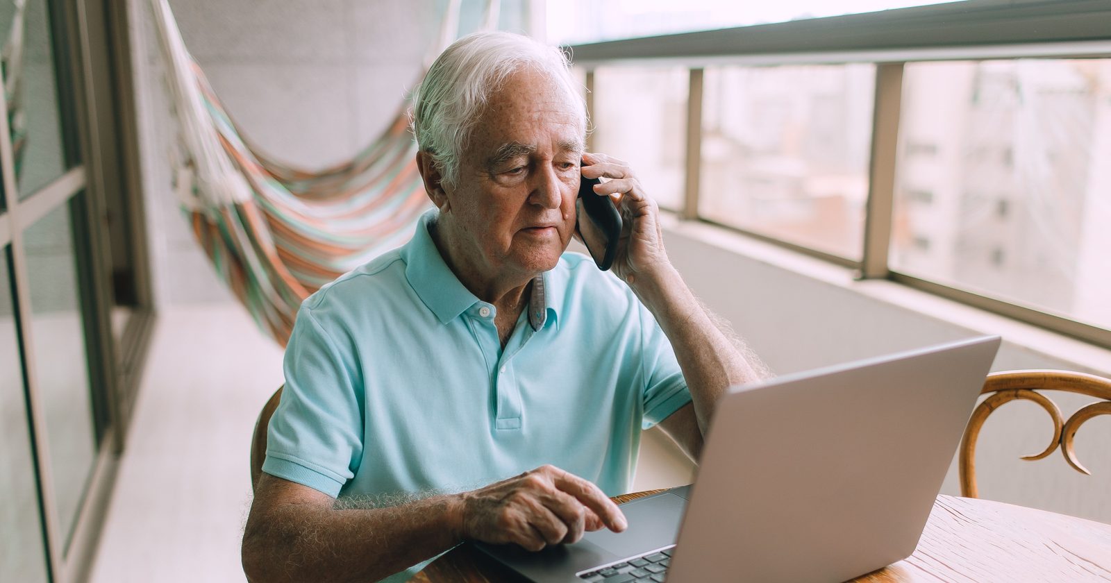 Elderly man multitasking on phone and laptop at home.