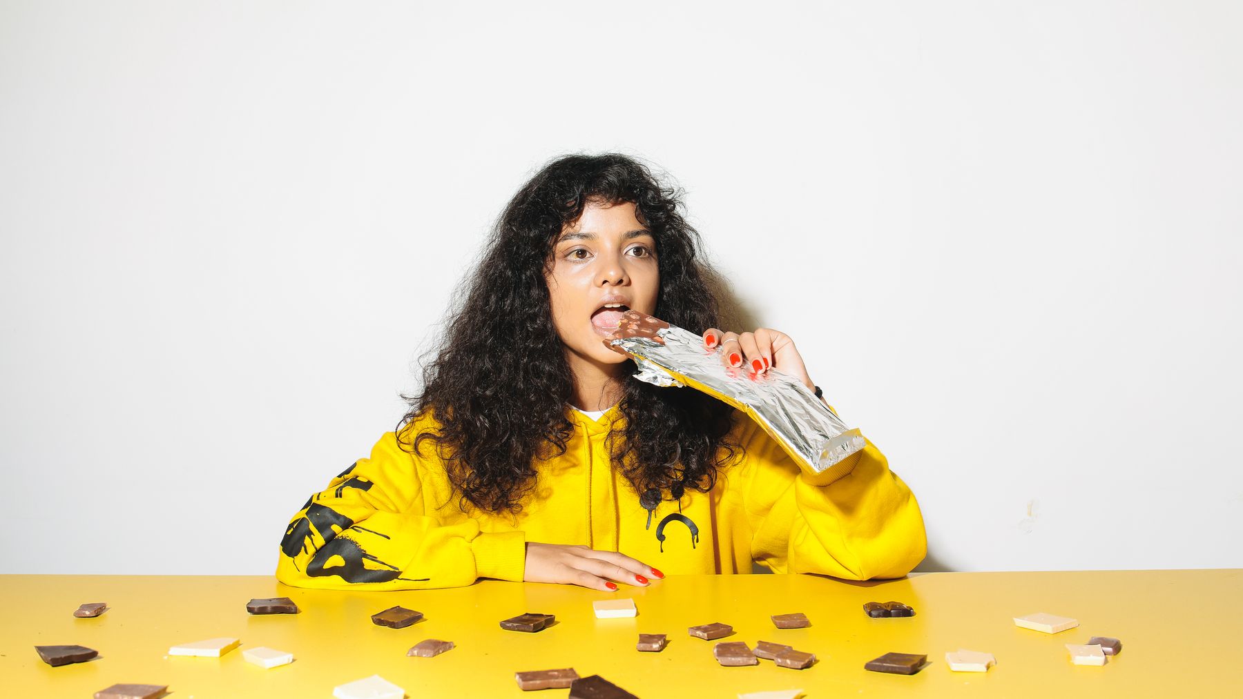Woman in a yellow sweatshirt holding a chocolate bar at a table scattered with chocolate pieces