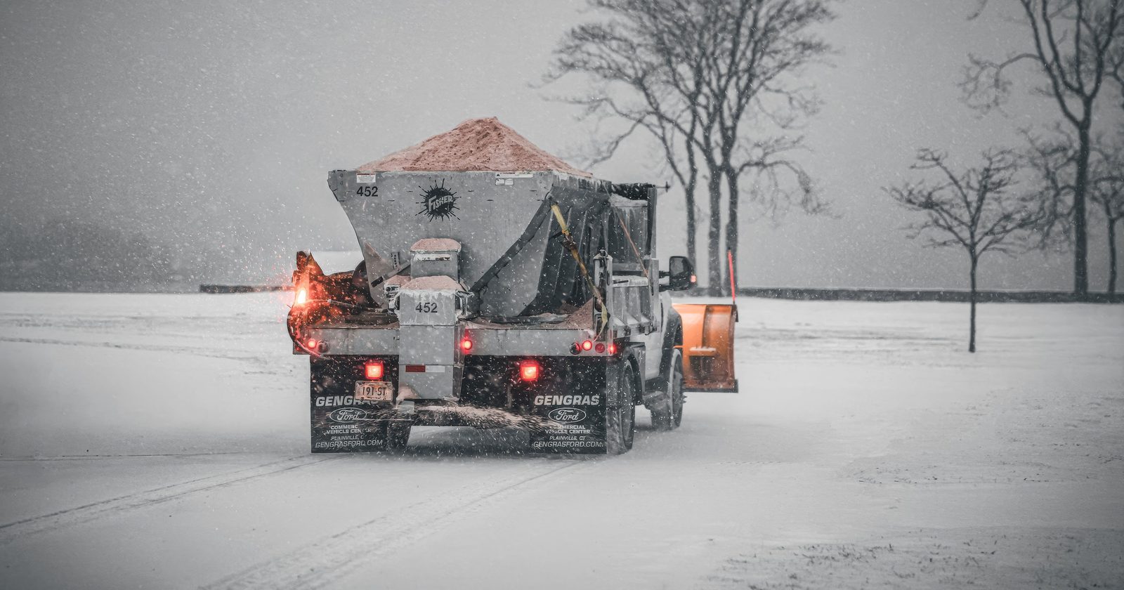 A snowplow truck spreading salt on a snowy road during a winter storm, with bare trees in the background.