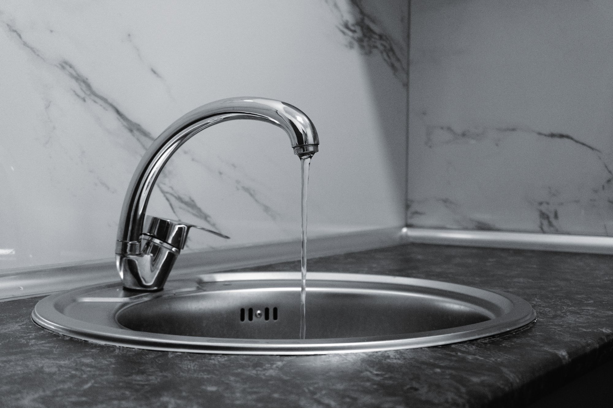 Water running from a chrome kitchen faucet into a round stainless steel sink, in black and white