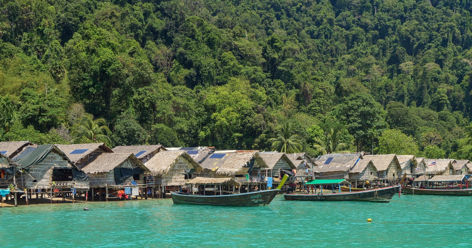 Thatched Moken stilt houses and long-tail boats over turquoise water below a forested hillside