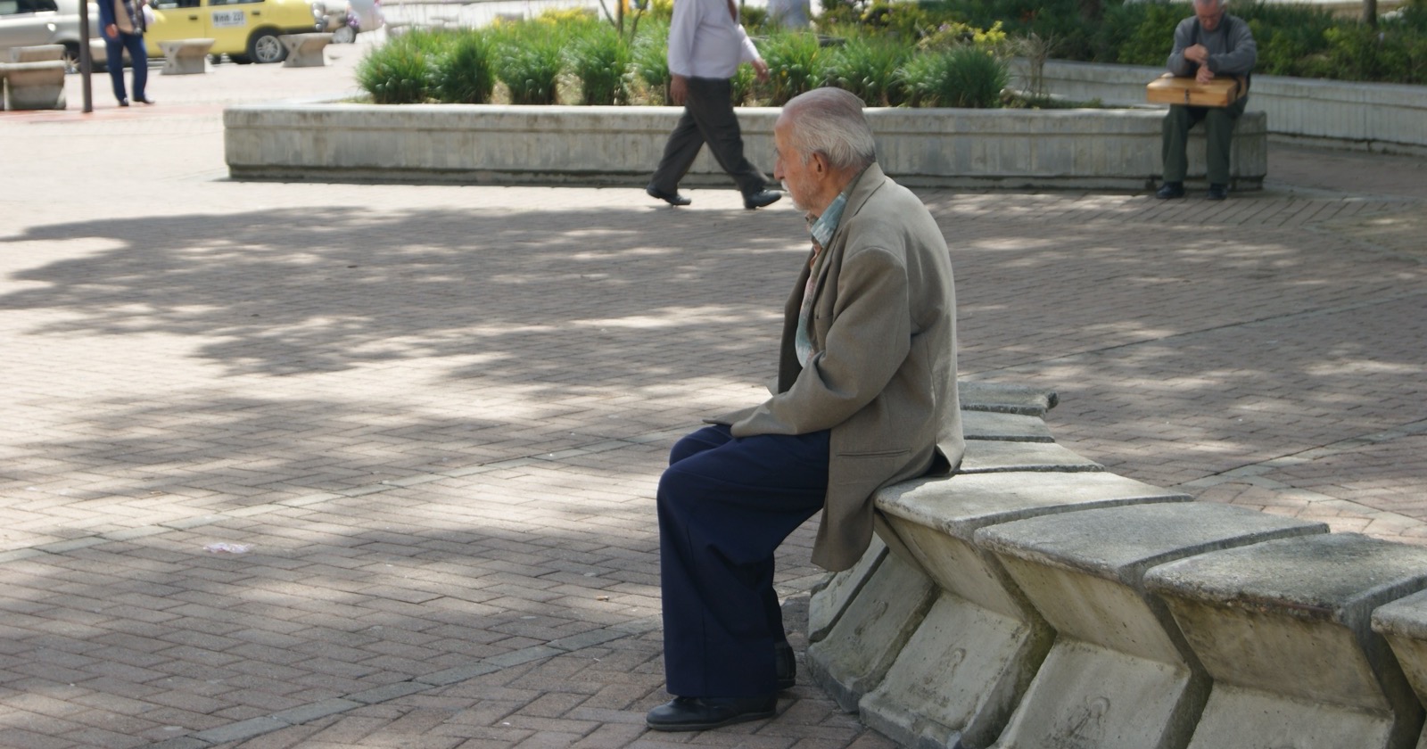 An older man sits alone on a stone bench in a city square as people walk past.