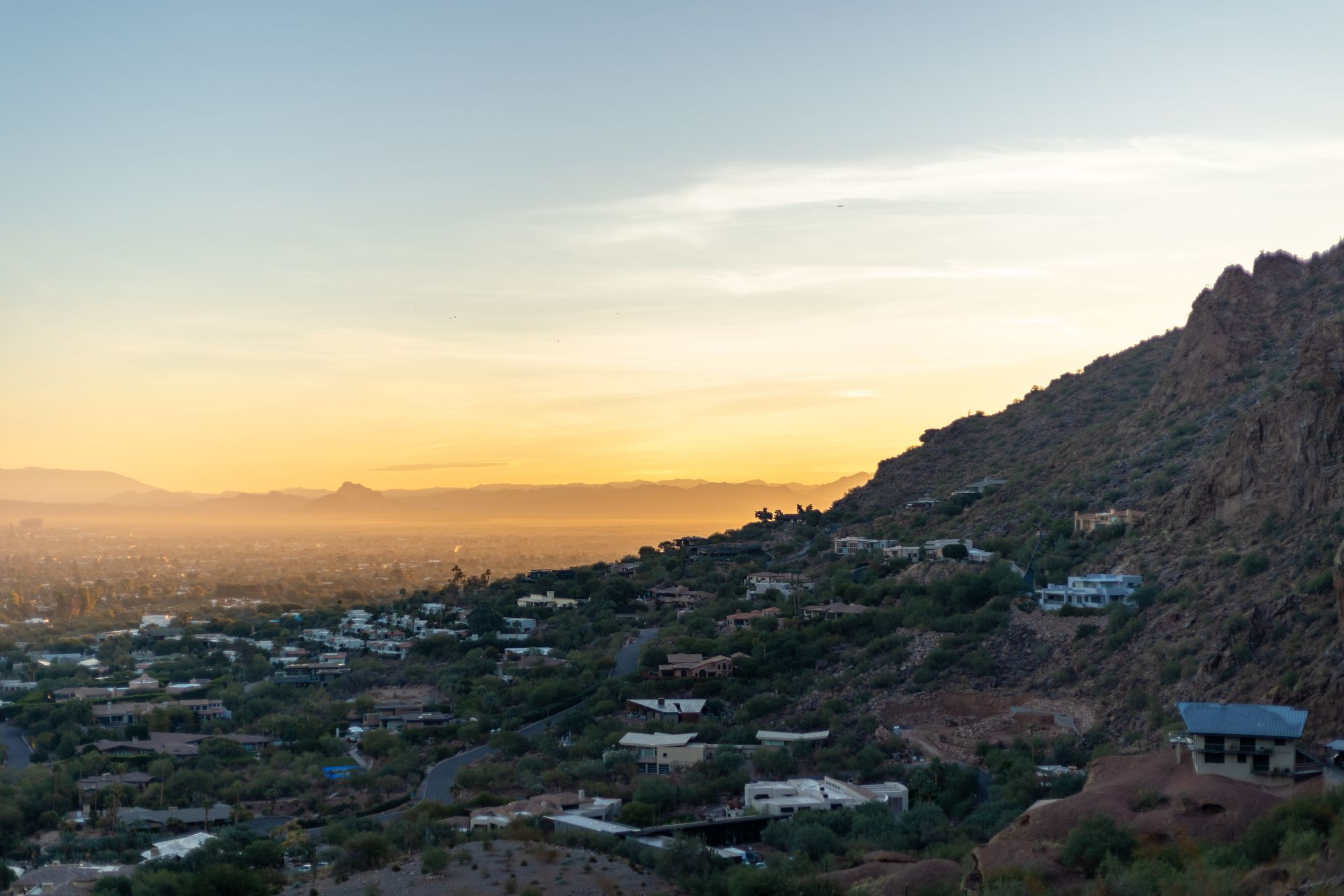 Hazy view over the Phoenix, Arizona, valley in low golden sunlight, with homes on a desert hillside in the foreground