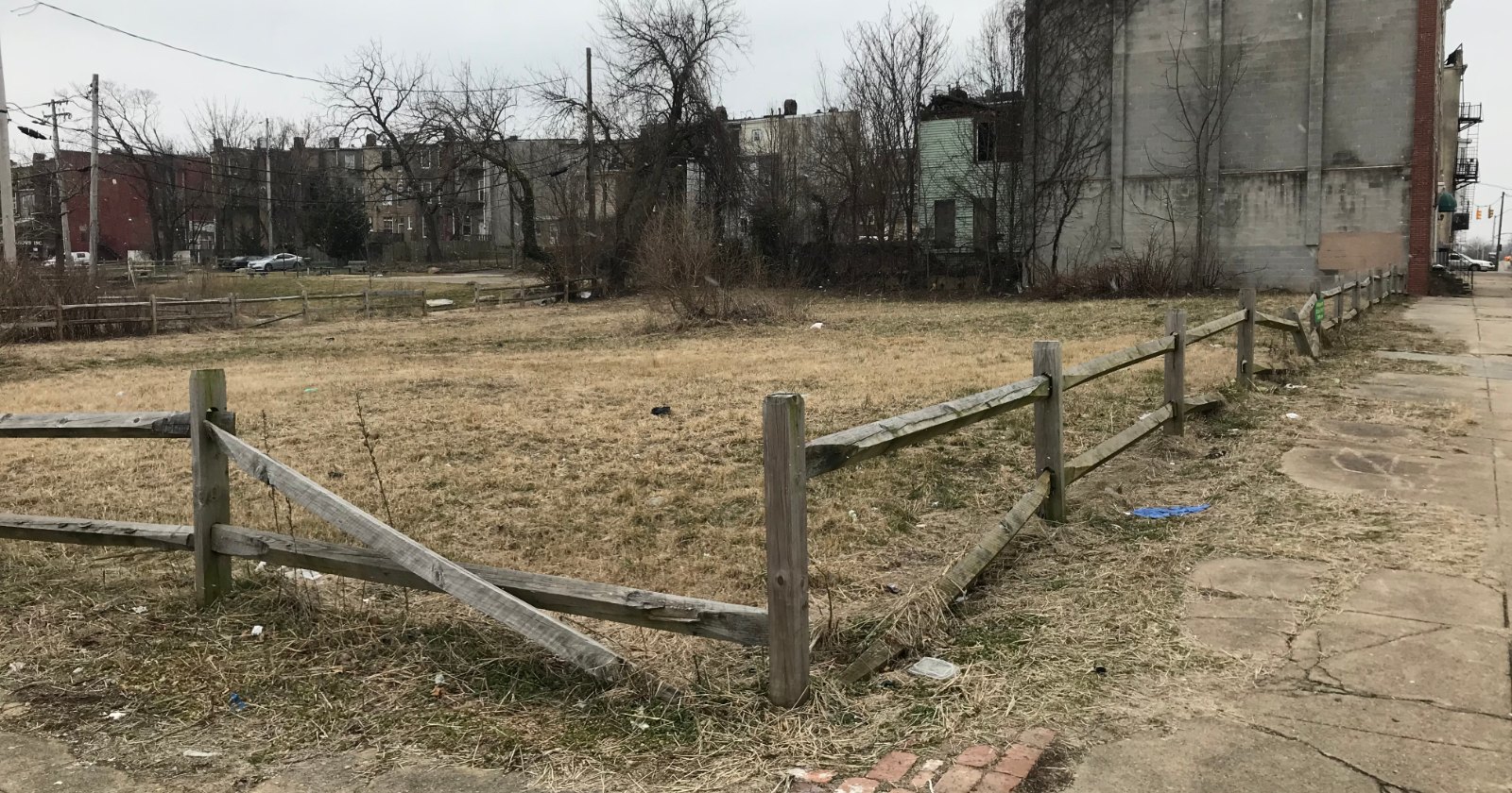 A grassy vacant lot edged by a low split-rail wooden fence, with rowhouses and bare trees behind it.